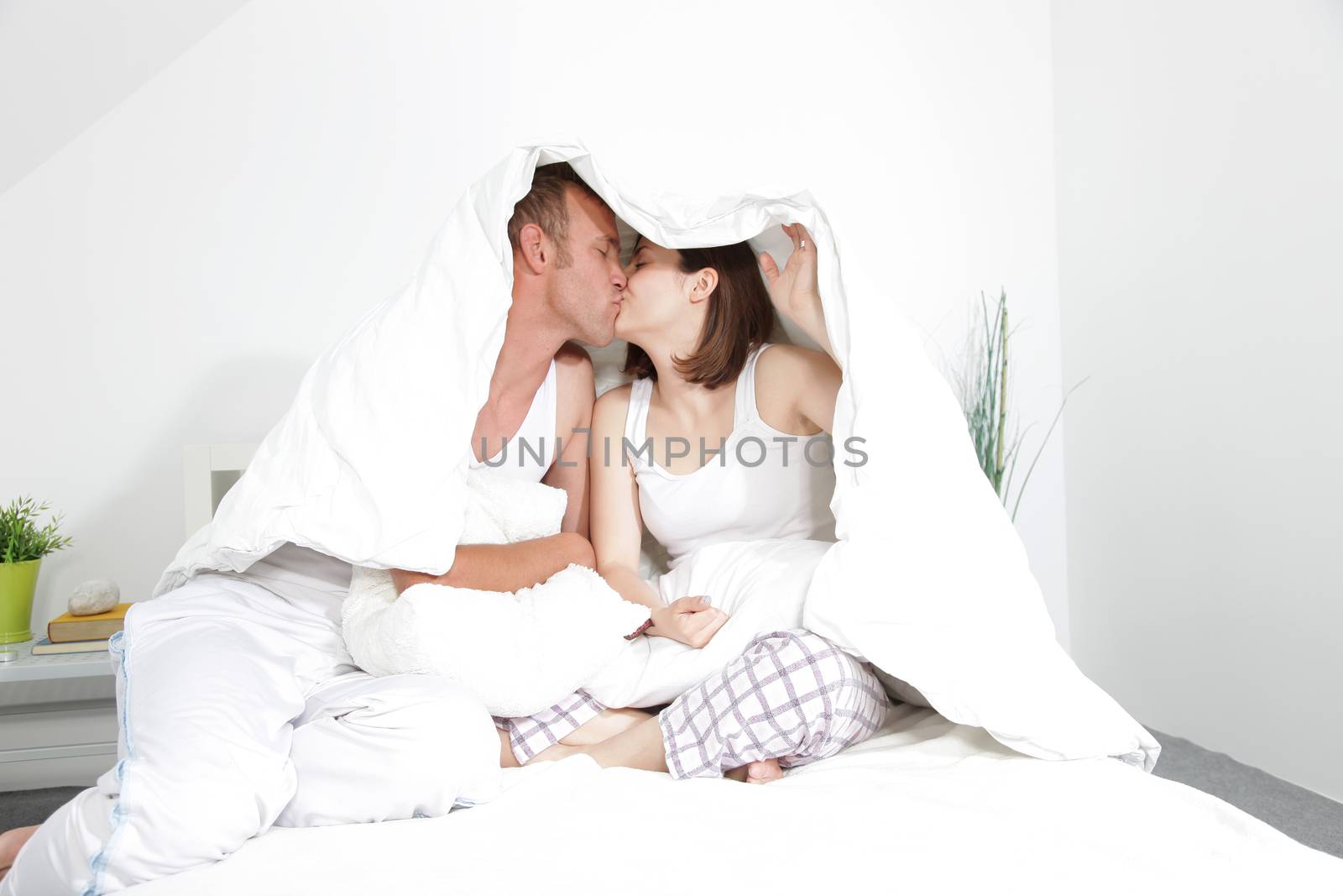 Loving couple kissing under the duvet sitting on their bed in the bedroom sharing an affectionate moment
