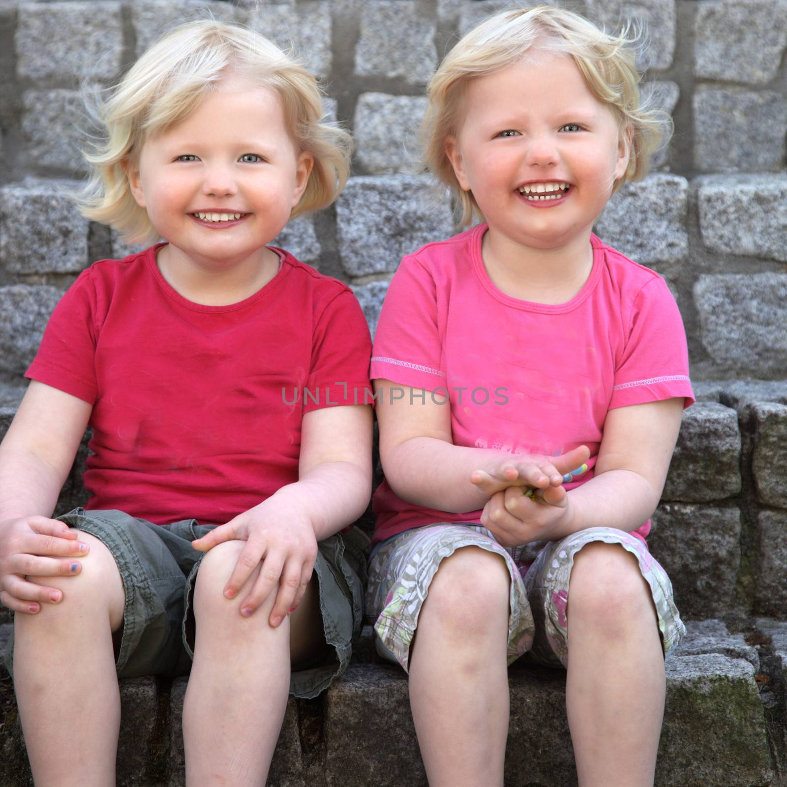 Laughing cute identical blond twins sitting against a stone wall looking at the camera with charming smiles