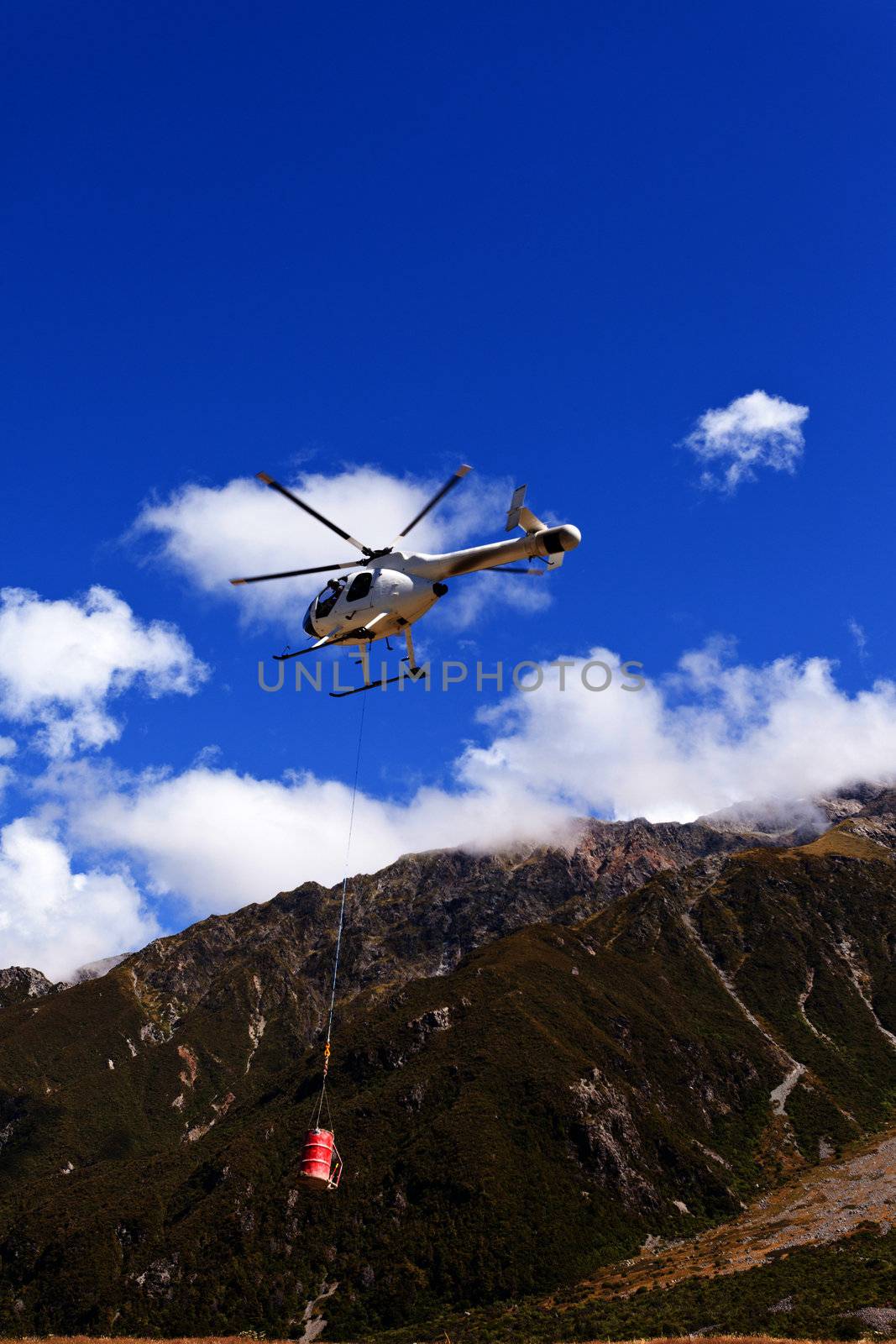 Small helicopter with transport cargo bucket flying in blue sky over mountains of Southern Alps New Zealand to supply remote site