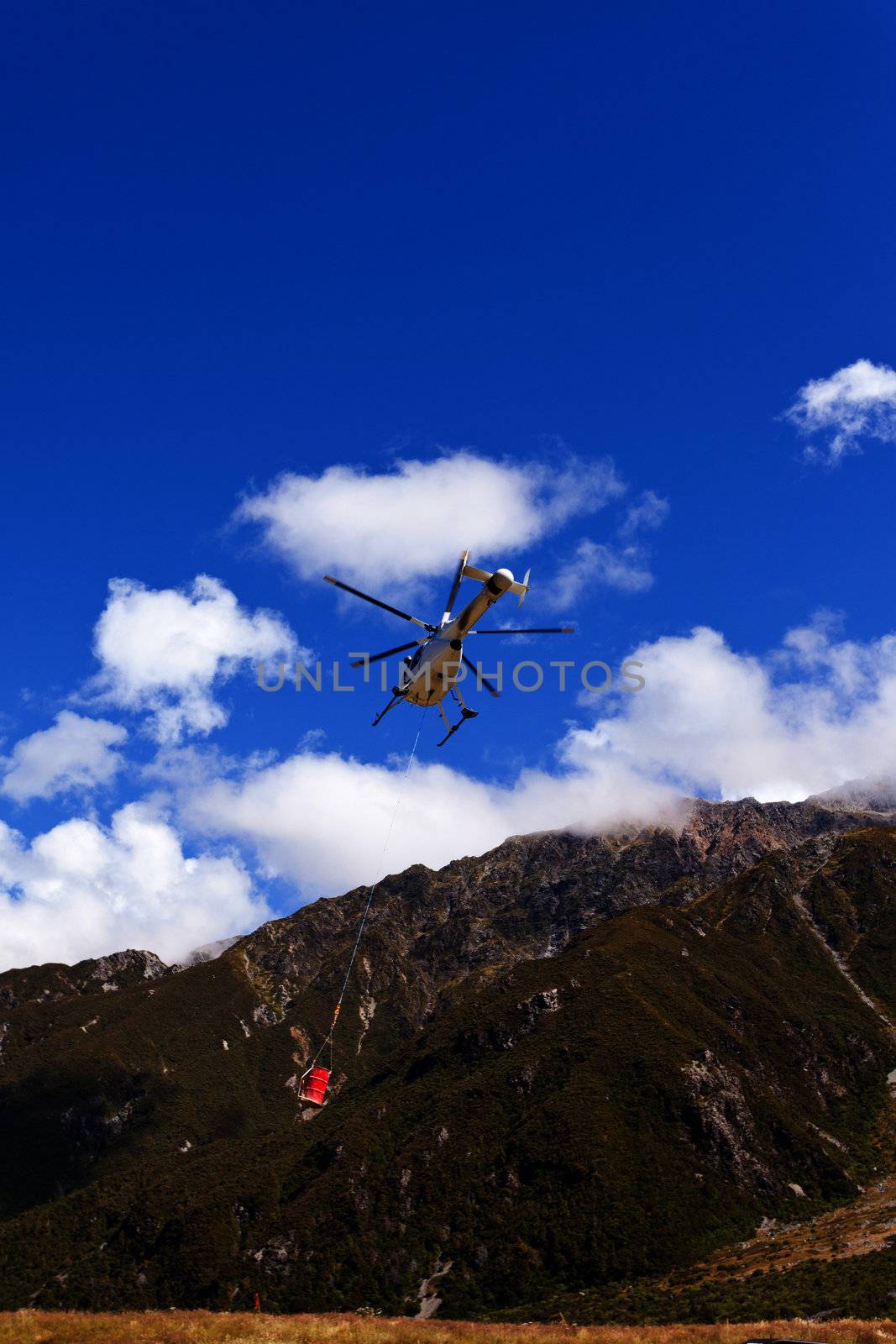 Small helicopter with transport cargo bucket flying in blue sky over mountains of Southern Alps New Zealand to supply remote site