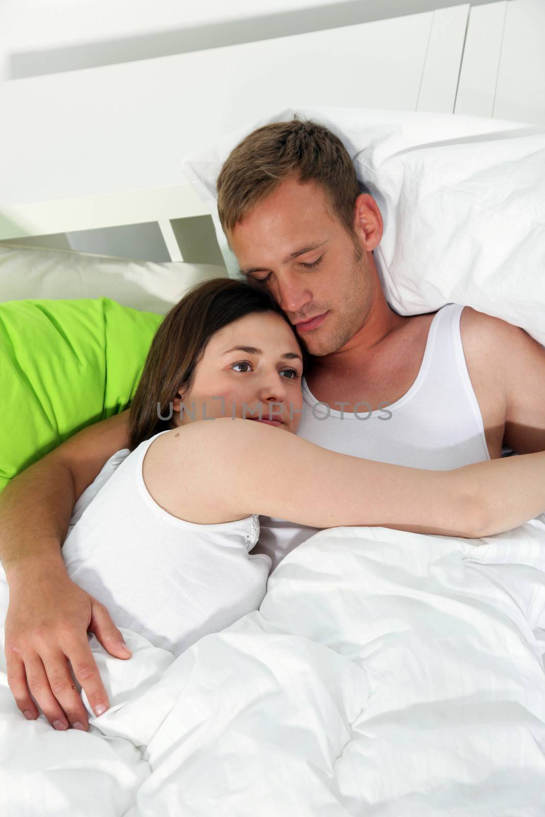 Young couple relaxing together in bed entwined in each others arms with the husband already falling asleep while his wife lies thinking