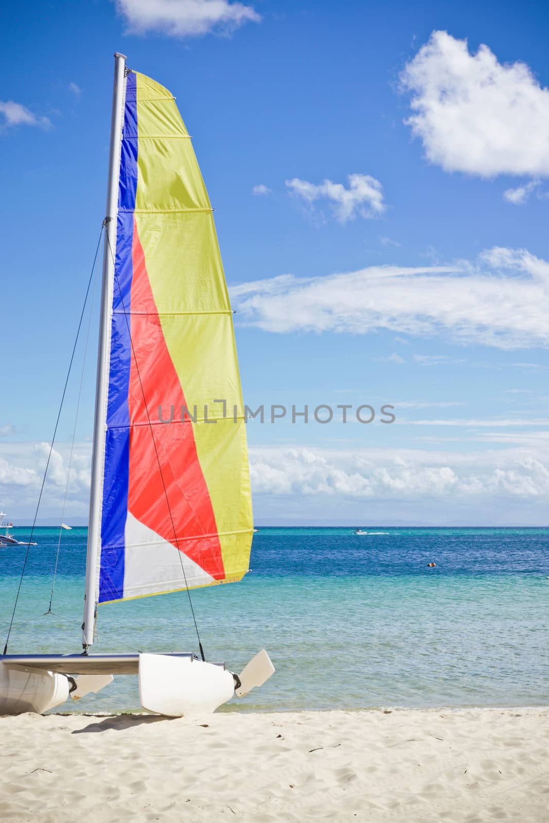 Small catamaran sailing dinghy on a sandy beach with a colourful sail against a tropical ocean backdrop