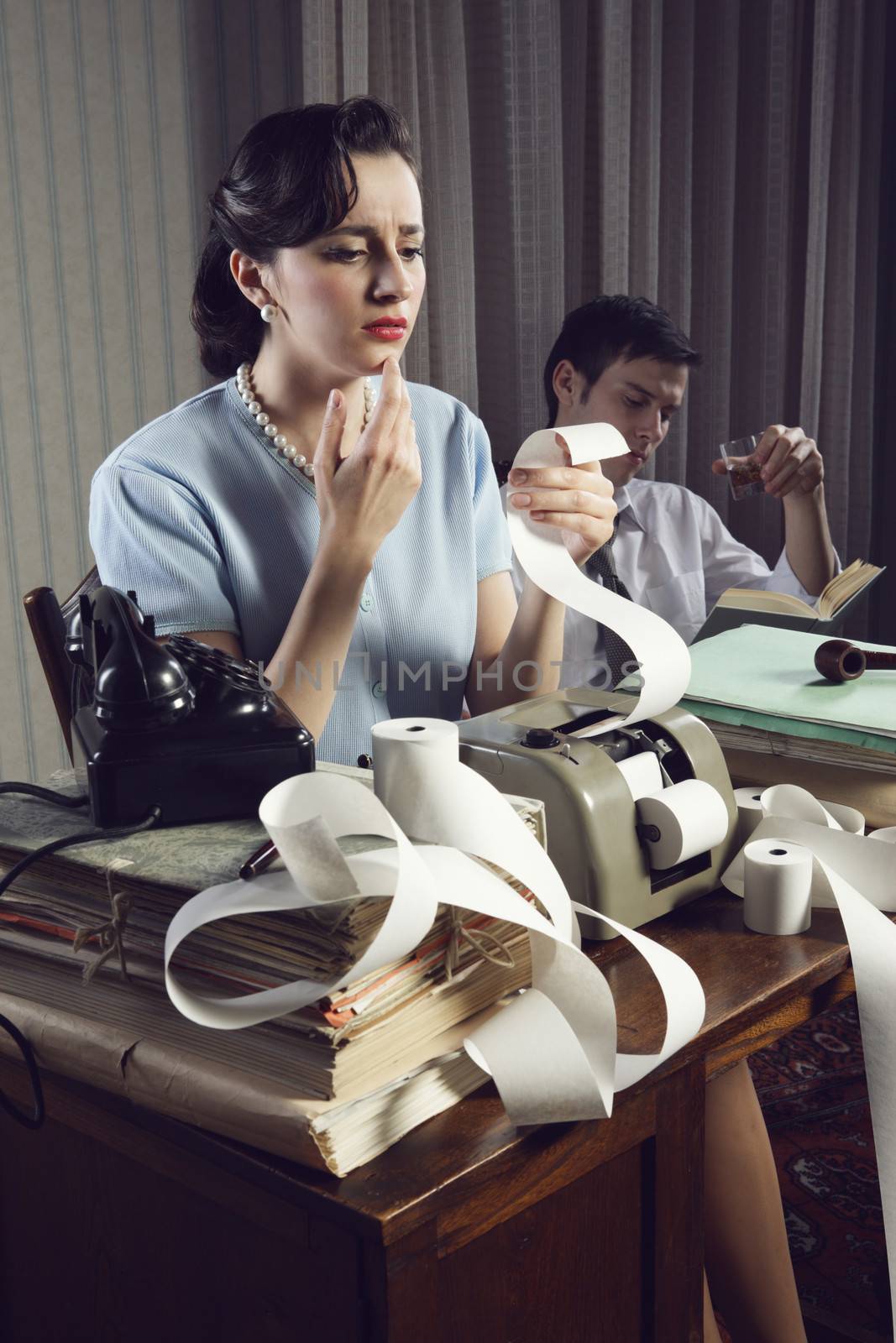 Woman works on home finances. Man relaxed in chair drinking whiskey adn reading a book