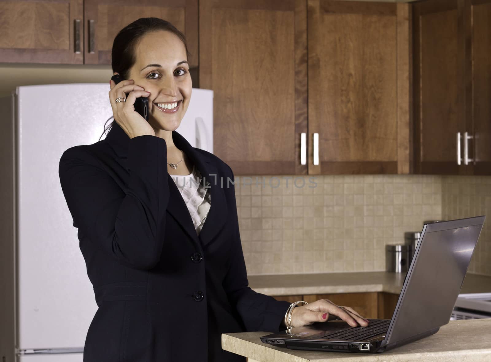 A business woman using a telephone and a laptop computer in her kitchen.