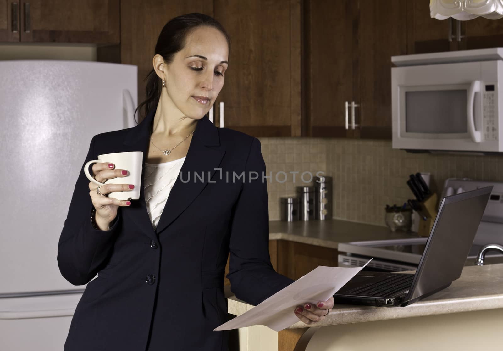 Business woman working on a laptop computer and drinking coffer in her kitchen.