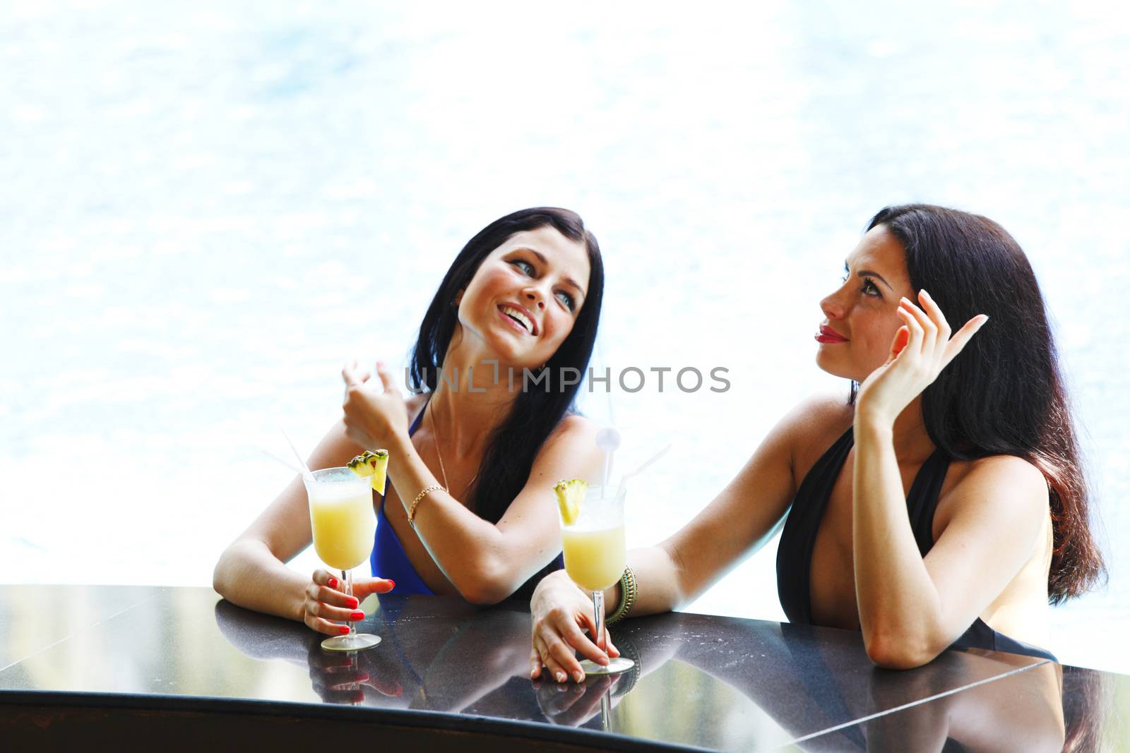 Two women with cocktails in swimming pool by Yellowj
