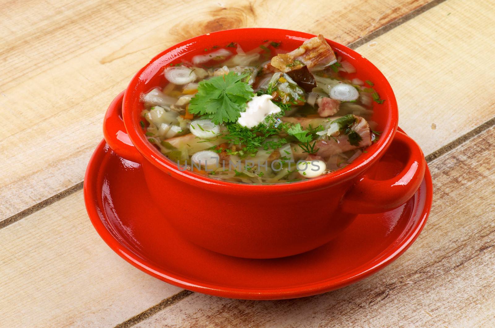 Delicious Vegetable Soup with Smoked Pork Ribs, Cabbage, Leek, Carrot, Garlic Greens and Sour Cream in Red Bowl isolated on Wooden background