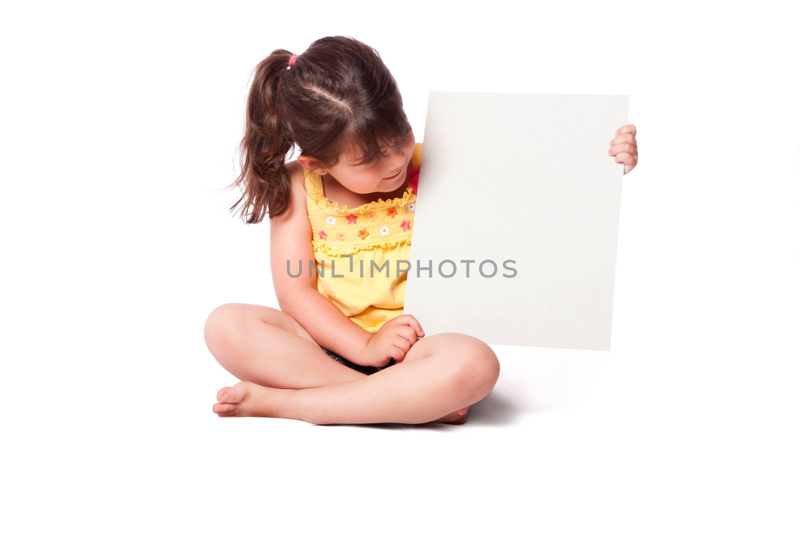 Cute girl wearing summery yellow shirt sitting while holding whiteboard and looking at it.