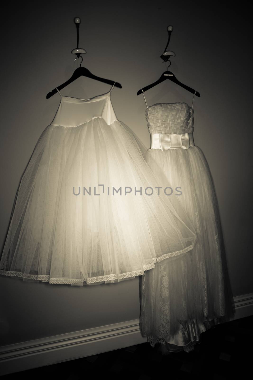 Two beautiful filmy wedding gowns with graceful full skirts hanging on clothes hangers on a wall in a darkened room