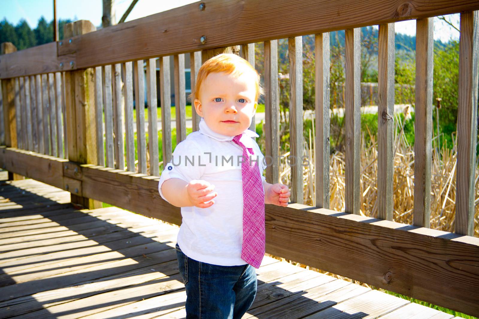 A one year old baby boy walks across a bridge wearing nice clothes and a necktie.