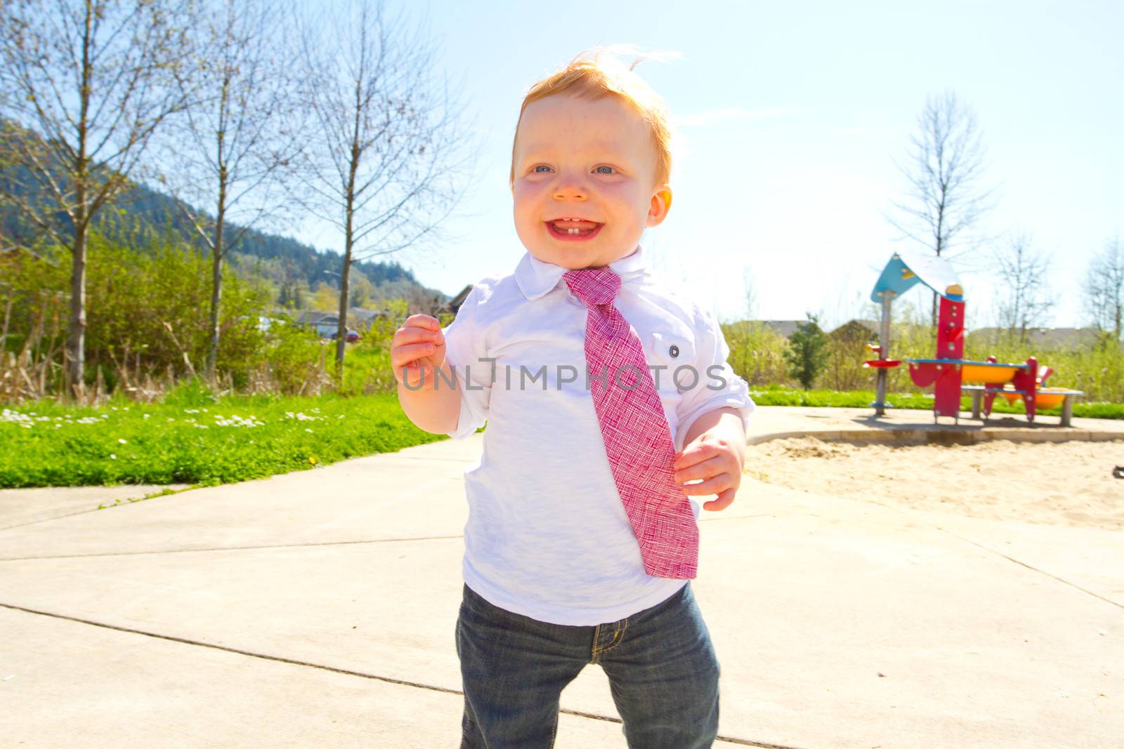 A baby boy plays at the park wearing his Sunday's best clothes including a tie around his neck.
