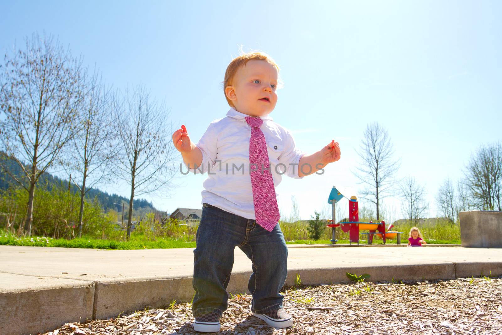 A baby boy plays at the park wearing his Sunday's best clothes including a tie around his neck.