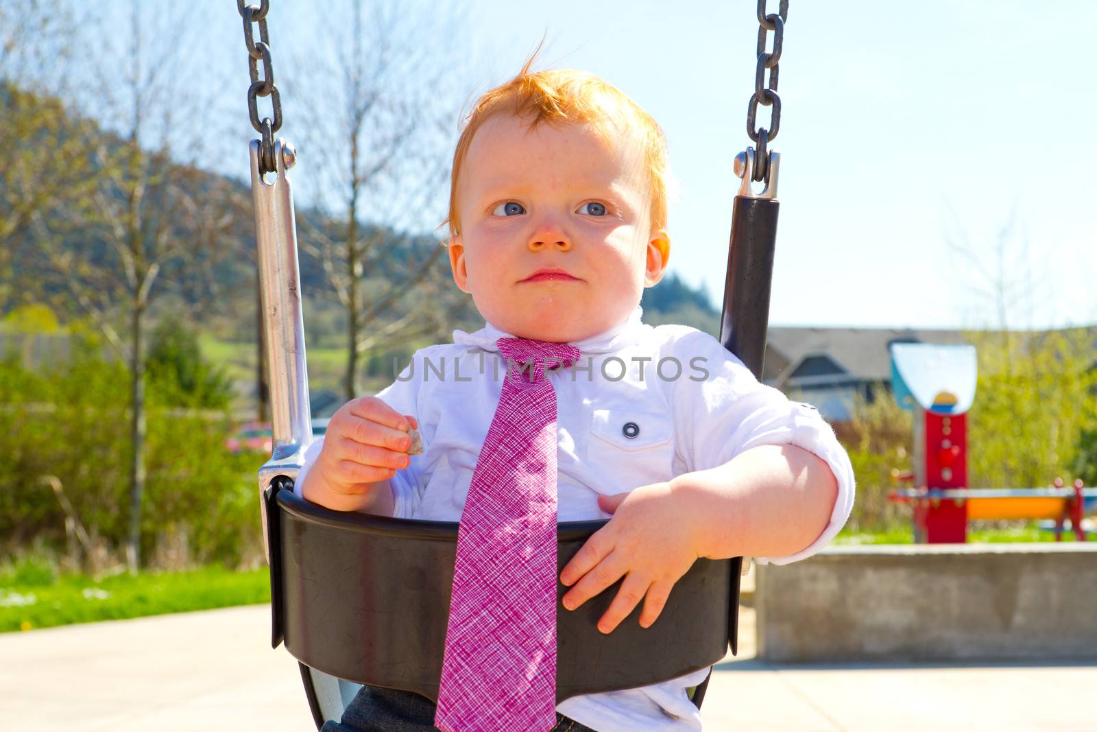 A baby boy plays on a swing set at the park wearing nice clothing.