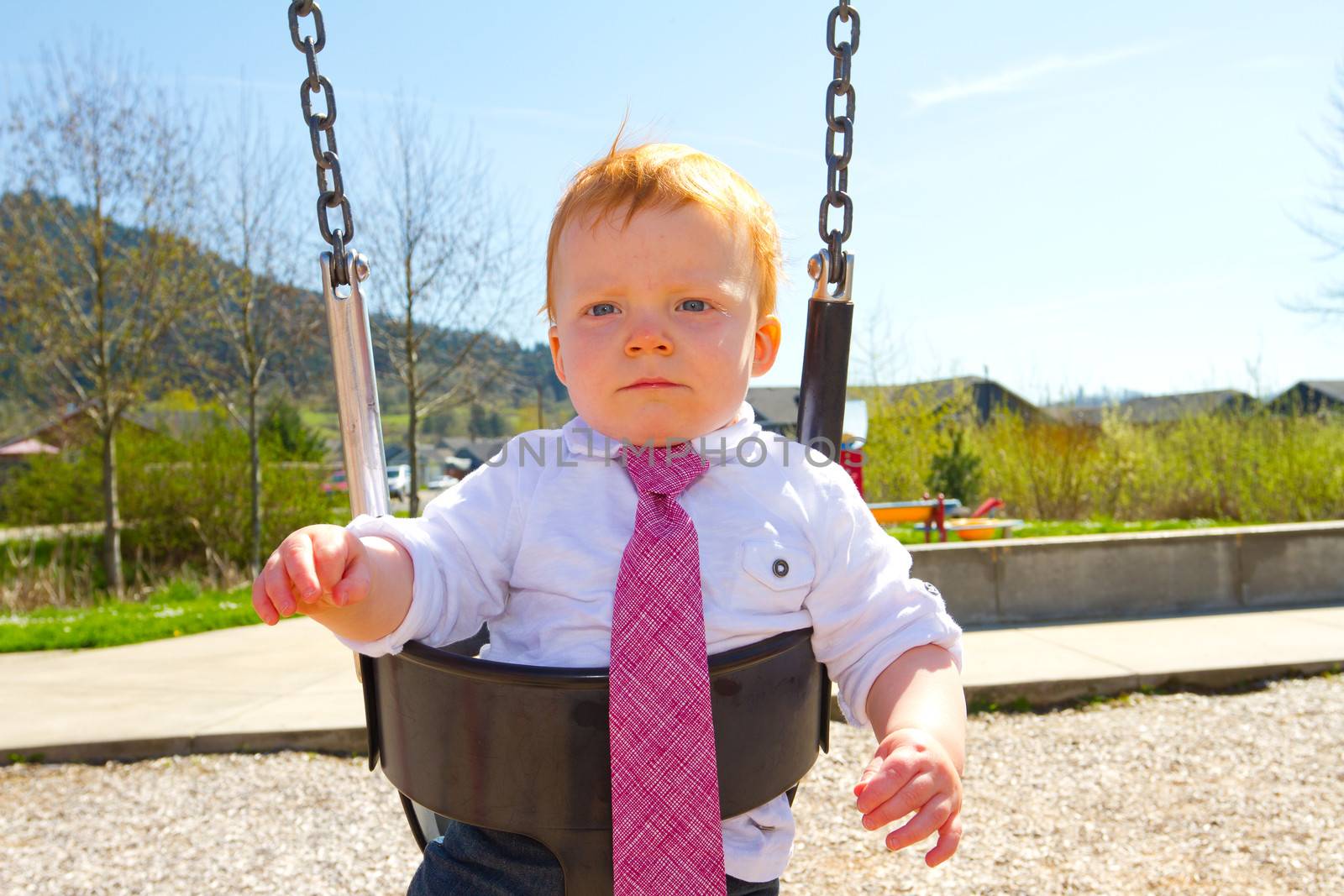 A baby boy plays on a swing set at the park wearing nice clothing.