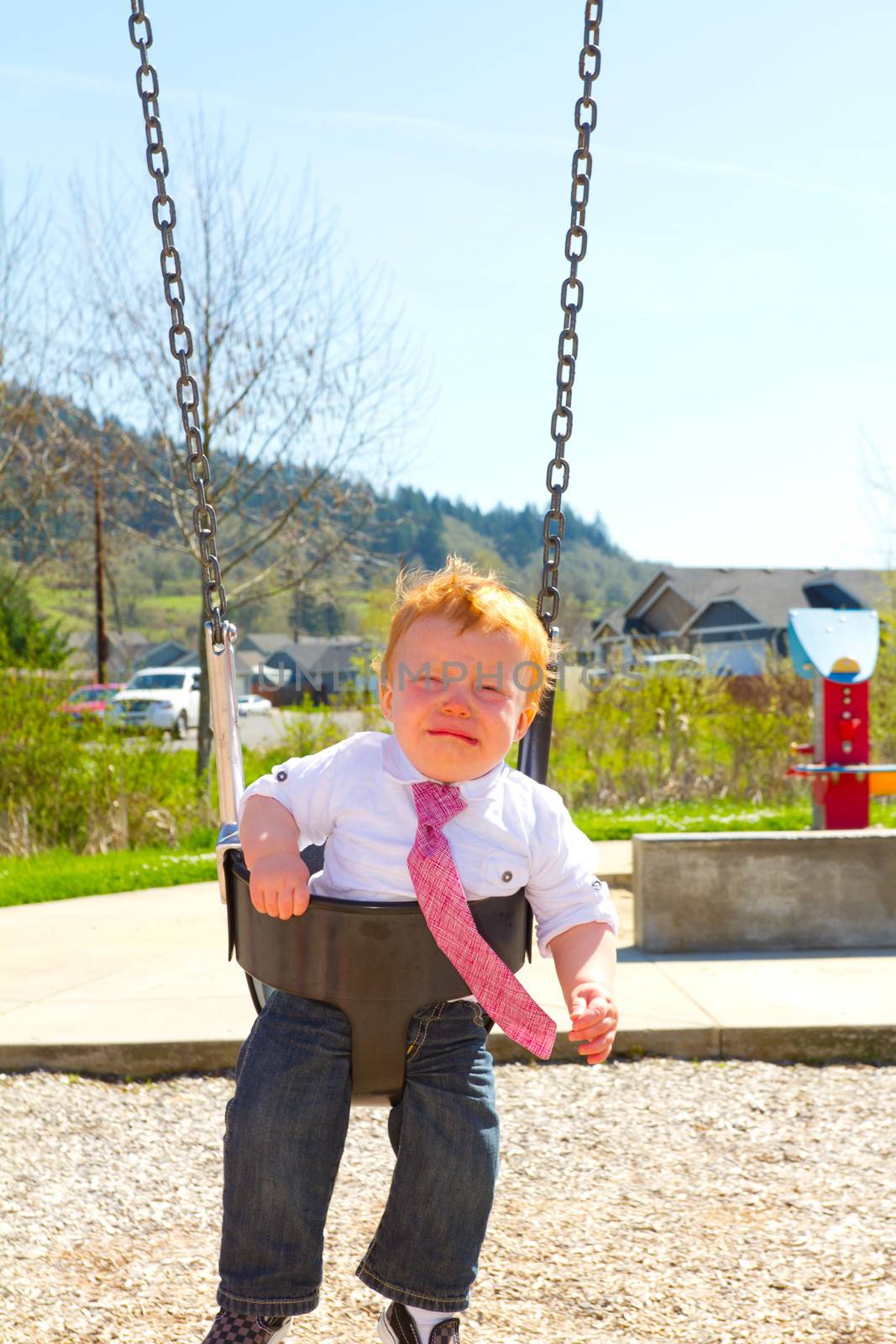 A baby boy sits on a swing set while upset and crying wearing nice clothes.