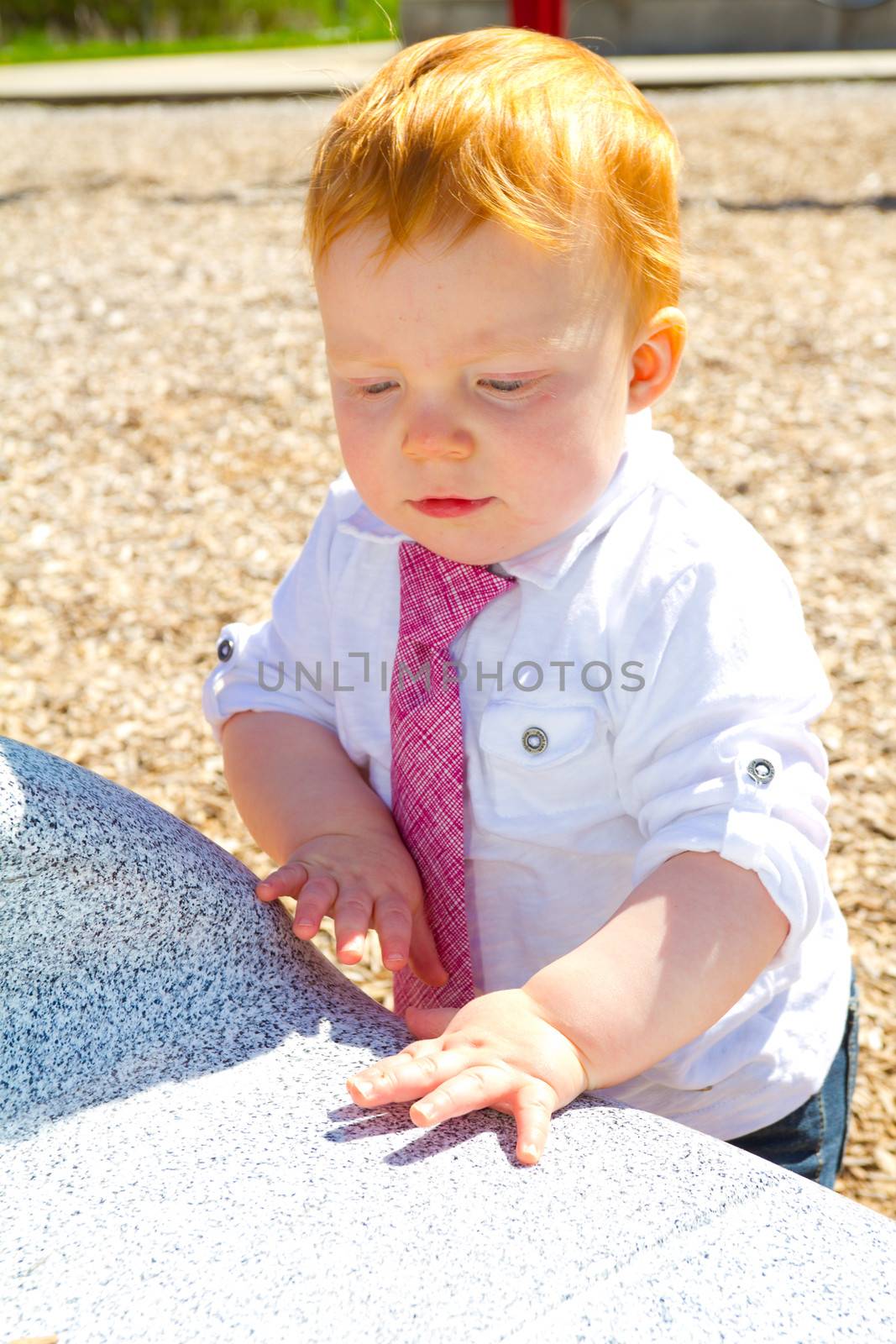 A caucasian baby boy plays at the park wearing a white shirt and a tie.