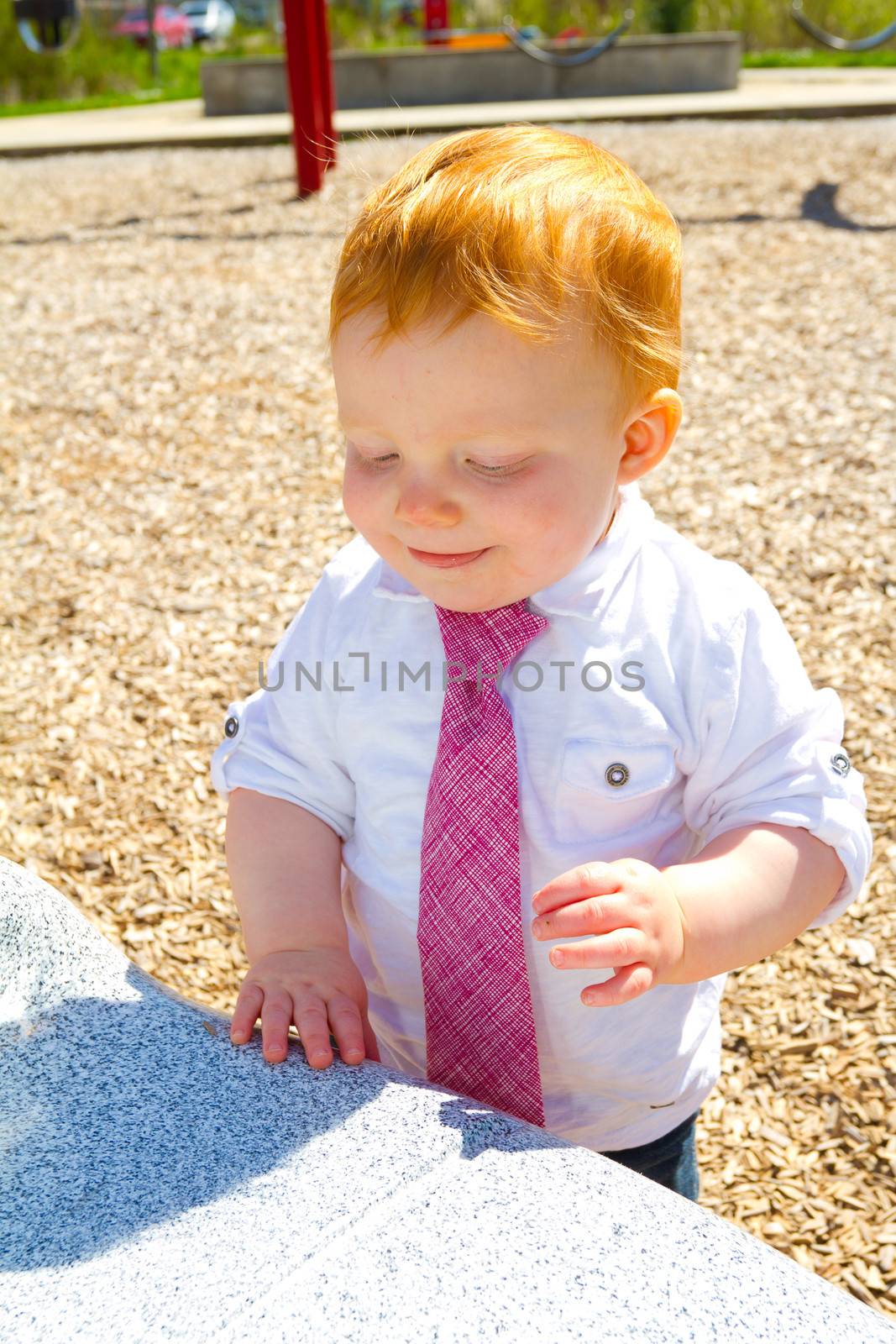 A caucasian baby boy plays at the park wearing a white shirt and a tie.