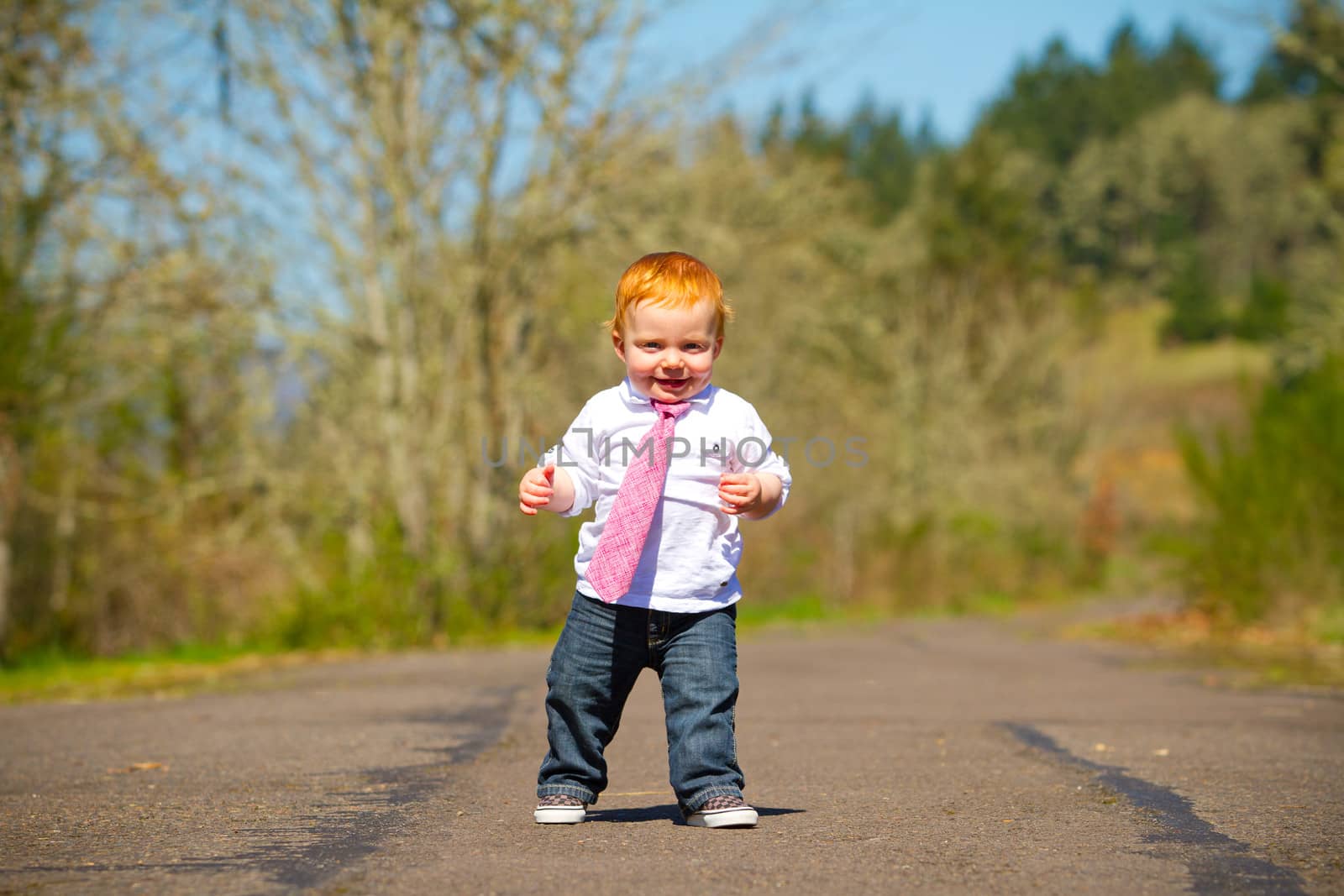 A one year old boy taking some of his first steps outdoors on a path with selective focus while wearing a nice shirt and a necktie.