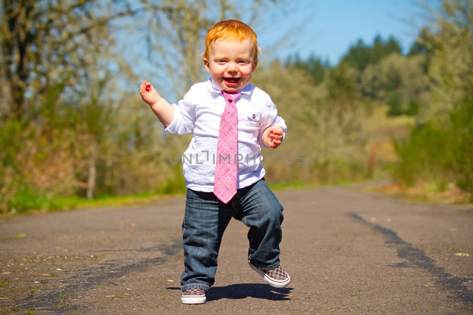 A one year old boy taking some of his first steps outdoors on a path with selective focus while wearing a nice shirt and a necktie.