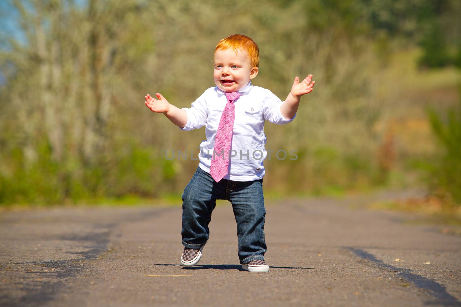 A one year old boy taking some of his first steps outdoors on a path with selective focus while wearing a nice shirt and a necktie.