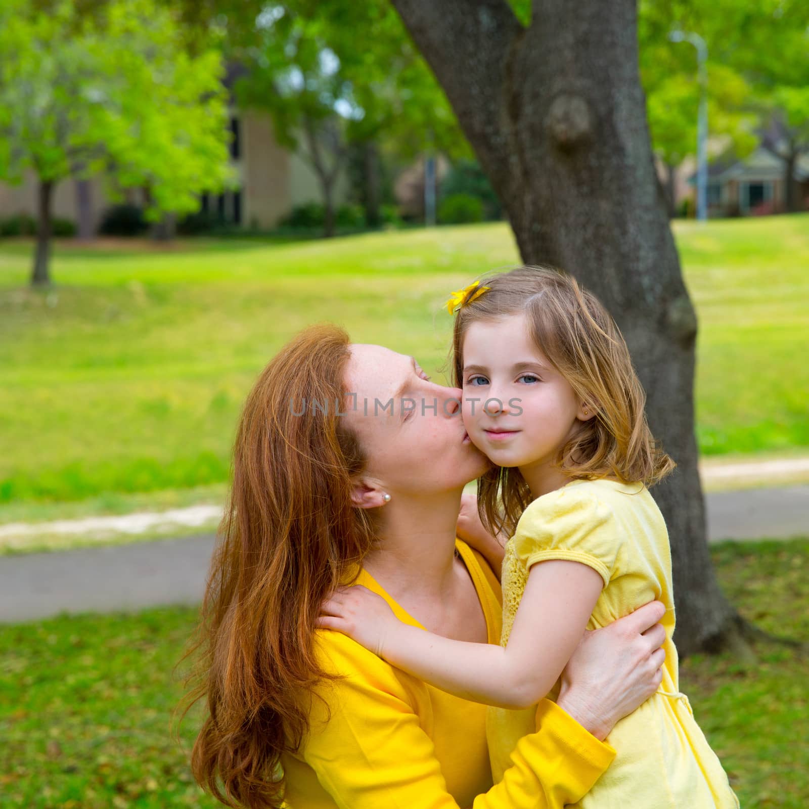 Mother kissing her blond daughter in green park outdoor dressed in yellow