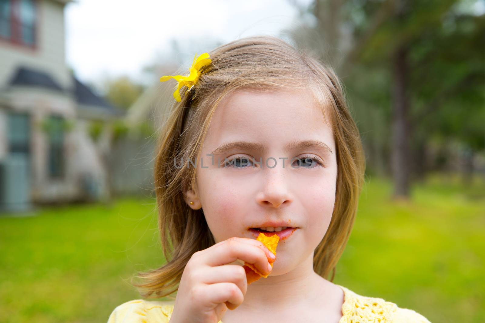 Blond kid girl eating corn snacks in outdoor park in lawn background