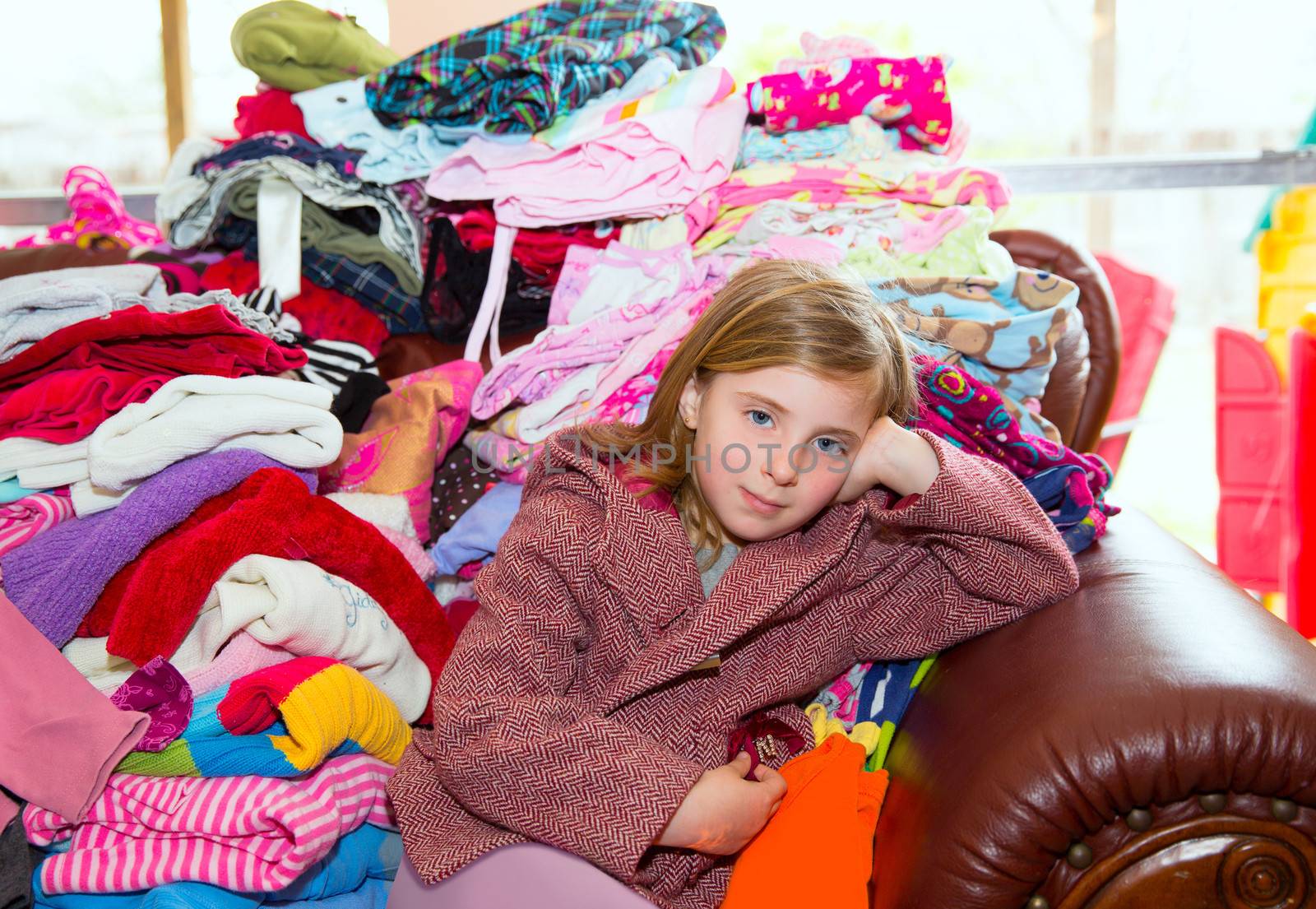 Blond kid girl sitting on a messy clothes sofa before folding laundry