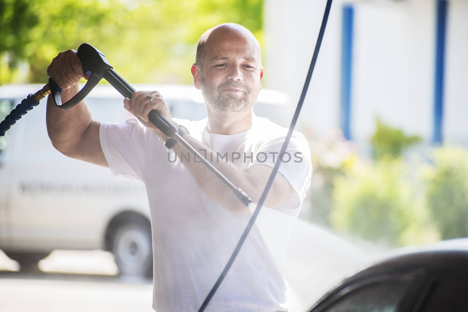 Bald man with a beard is washed with a pressure washer on a sunny day