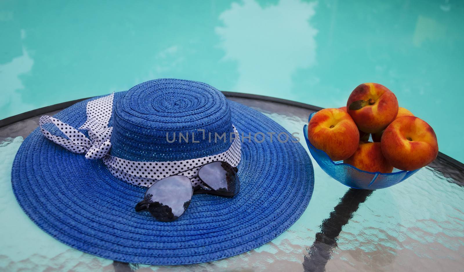 Woman's hat, sunglasses and peaches in glass plate are on table by the swimming pool