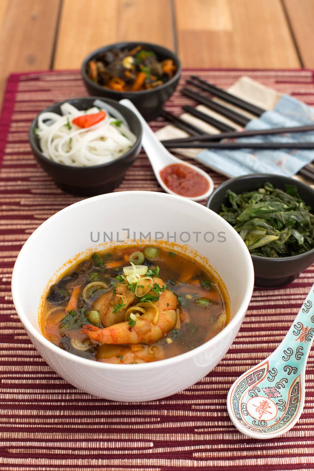 Asian soup with shrimps in white ceramic bowl composed with ceramic spoon with spicy red sauce, asian chopsticks and black bowls with rice noodles, kale (green cabbage) and fried vegetables. Composition on a old styled wooden table.