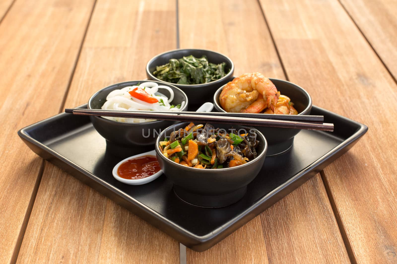 Traditional chinese dish on a square plate in black bowls with shrimp, rice noodles, kale (green cabbage) and fried vegetables. Composed with ceramic spoon with spicy red sauce and chinese chopsticks. Composition on a old styled wooden table.