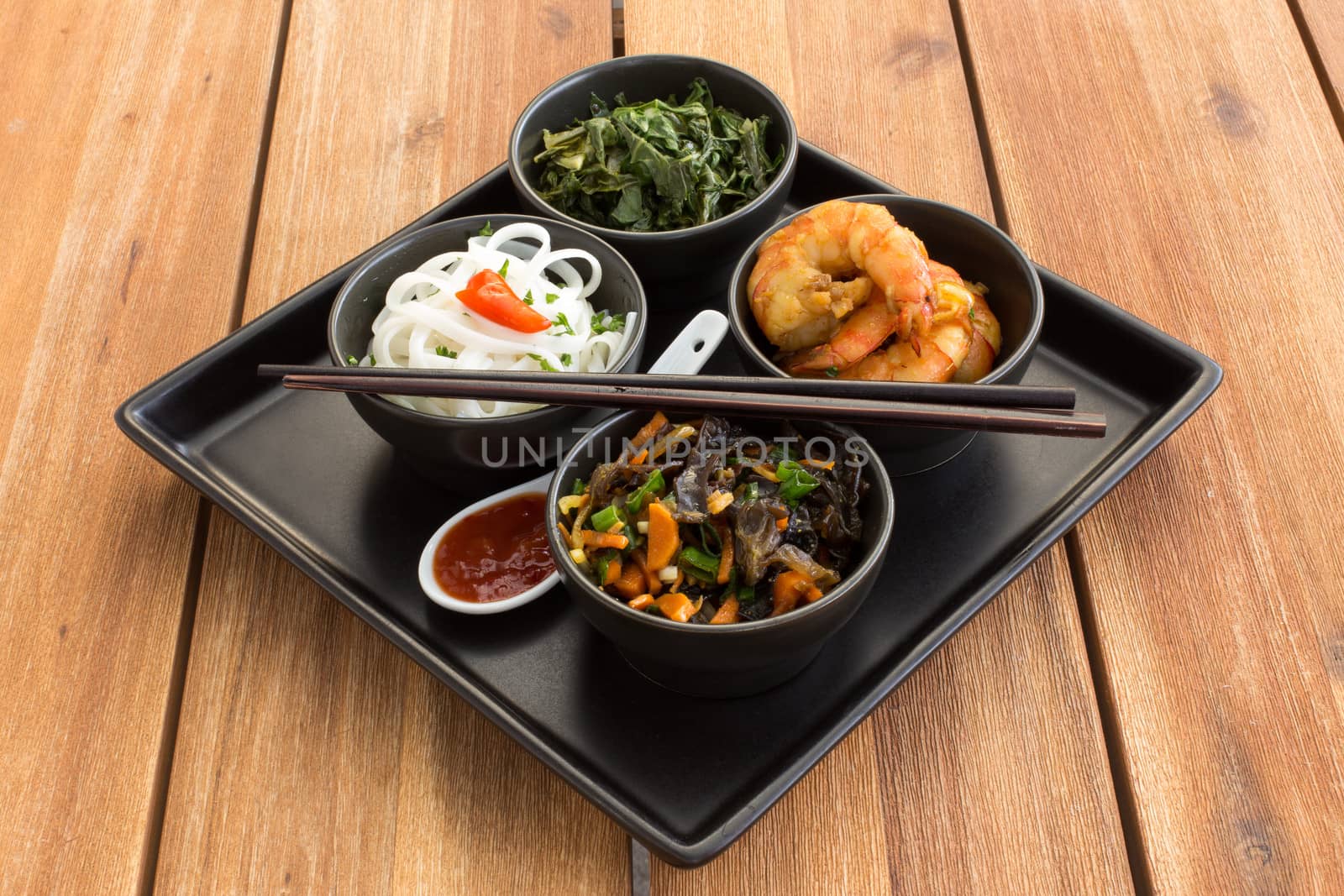 Traditional japanese dish on a square plate in black bowls with shrimp, rice noodles, kale (green cabbage) and fried vegetables. Composed with ceramic spoon with spicy red sauce and chinese chopsticks. Composition on a old styled wooden table.