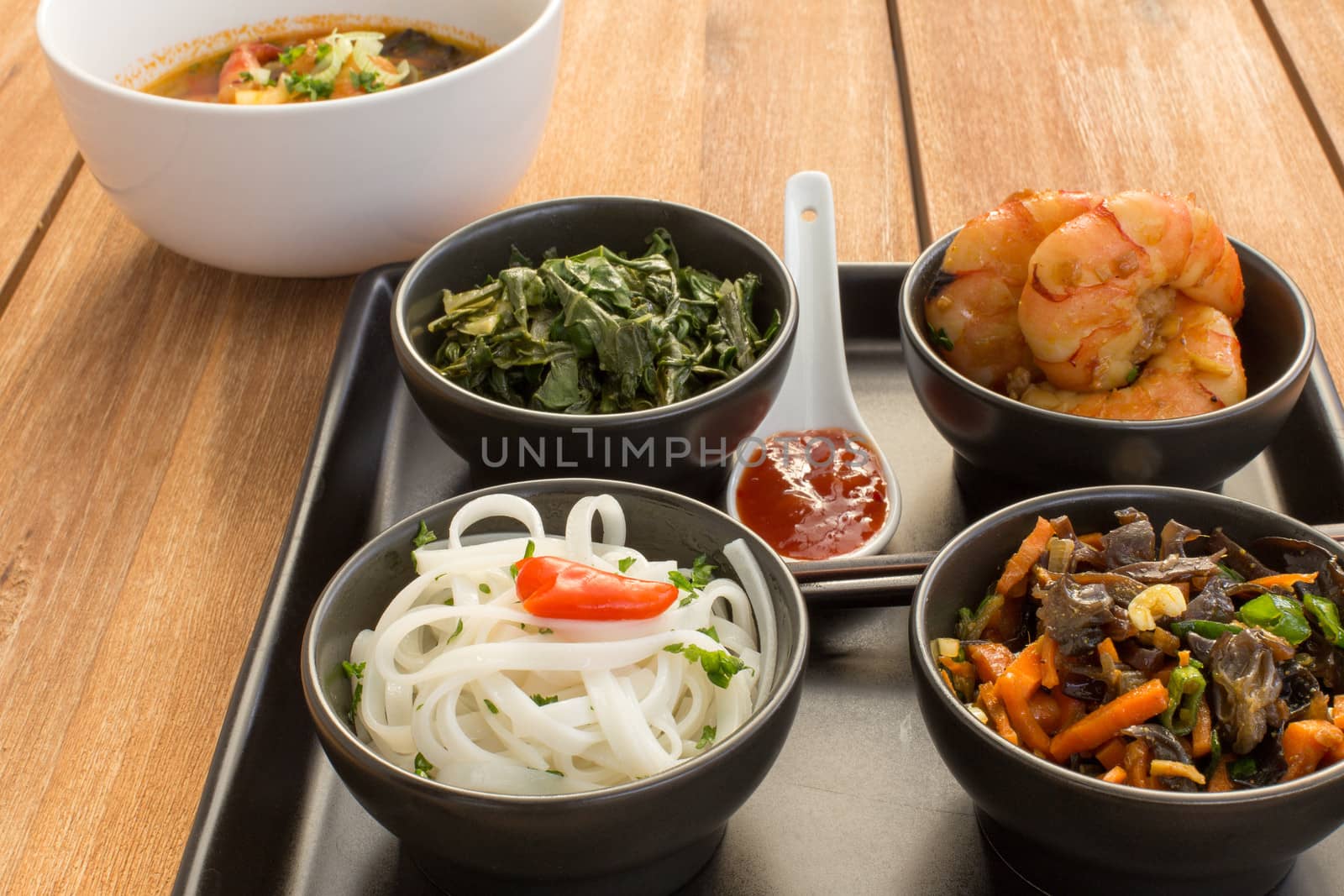 Asian dish on a square plate in black bowls with shrimp, rice noodles, kale (green cabbage) and fried vegetables. Composed with ceramic spoon with spicy red sauce, japanese chopsticks and a bowl of asian soup. Composition on a old wooden table.