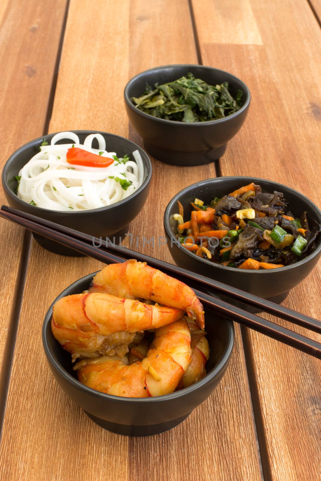 Asian vegetarian food composed with four black bowls with shrimps, rice noodles, kale (green cabbage), fried vegetables and chinese chopsticks. Composition on a old styled wooden table.