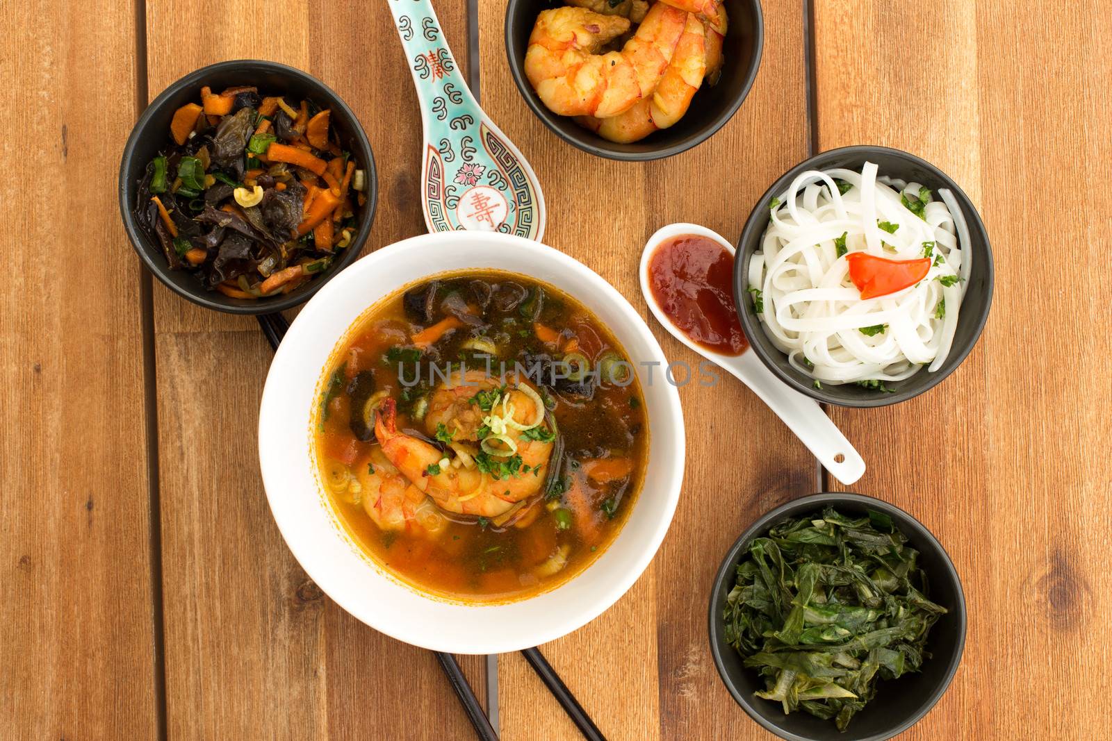 Asian vegetarian composition of food composed with four bowls of different appetizers (rice noodles, kale, fried vegetables and shrimps) and two ceramic spoons with asian sauce. Composition on a old styled wooden table.