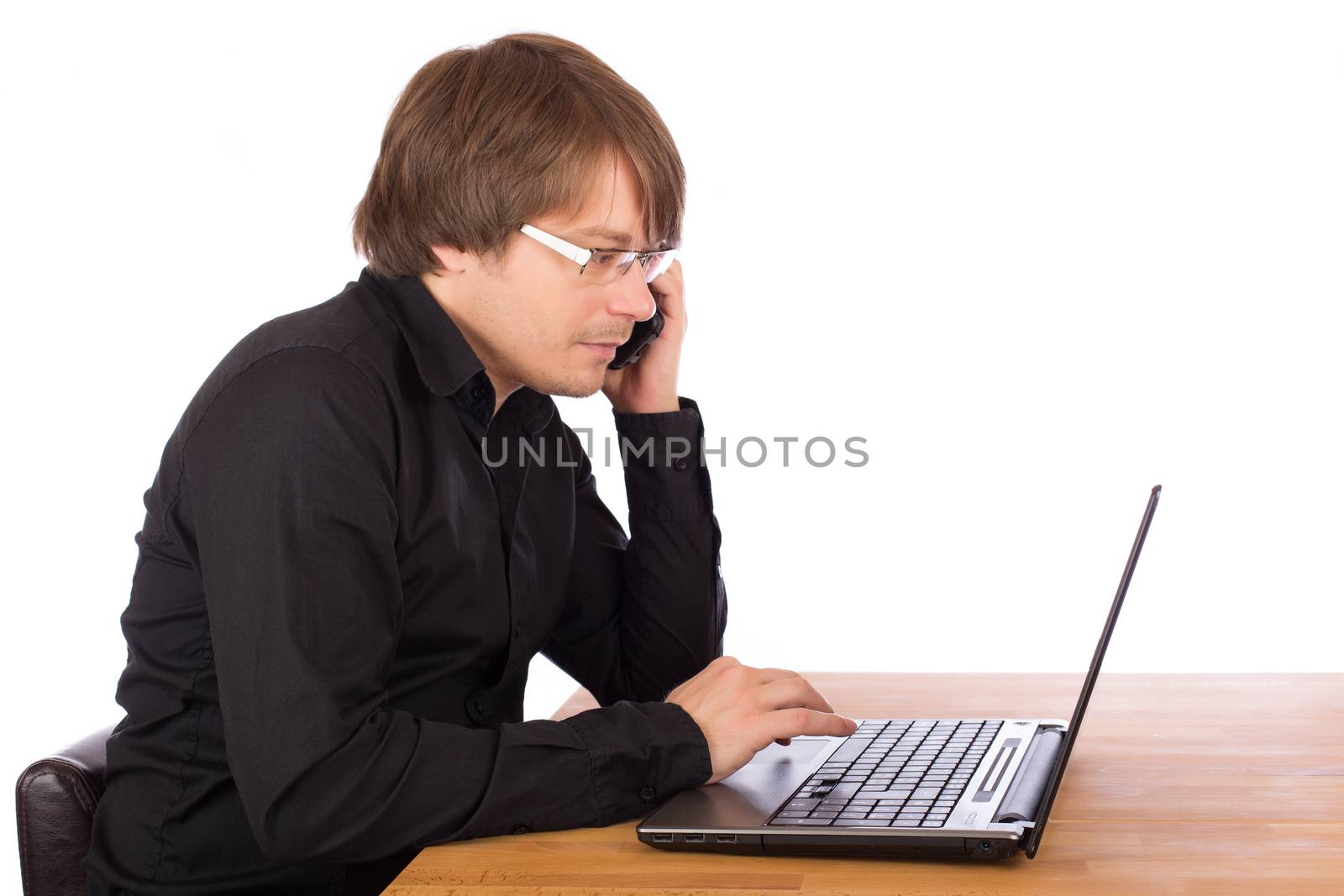 Young business man with black shirt and mobile phone working seriously on a laptop. Isolated on white background.