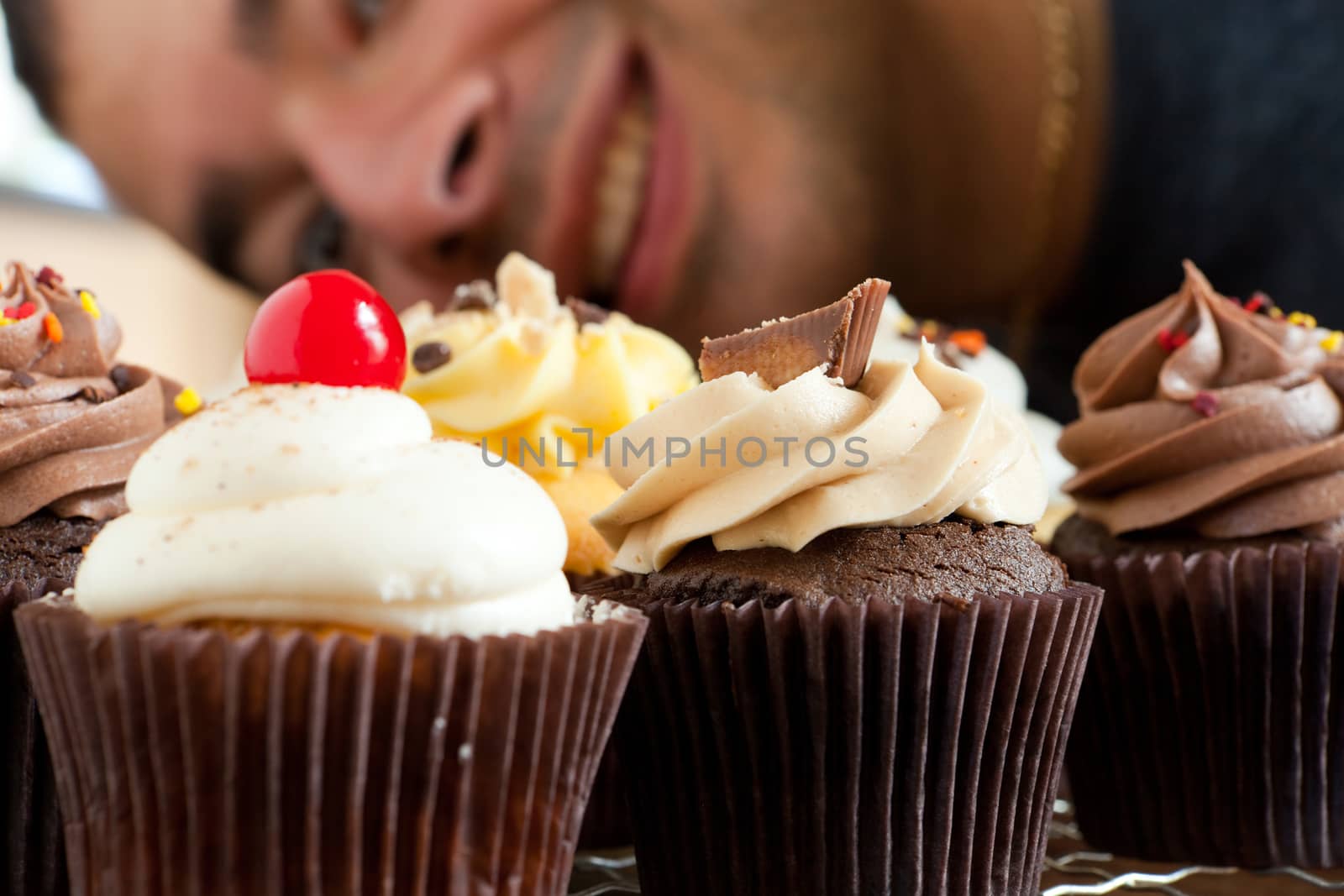 Close up of some decadent gourmet cupcakes frosted with a variety of frosting flavors.  Shallow depth of field with the face of a smiling man in the background.