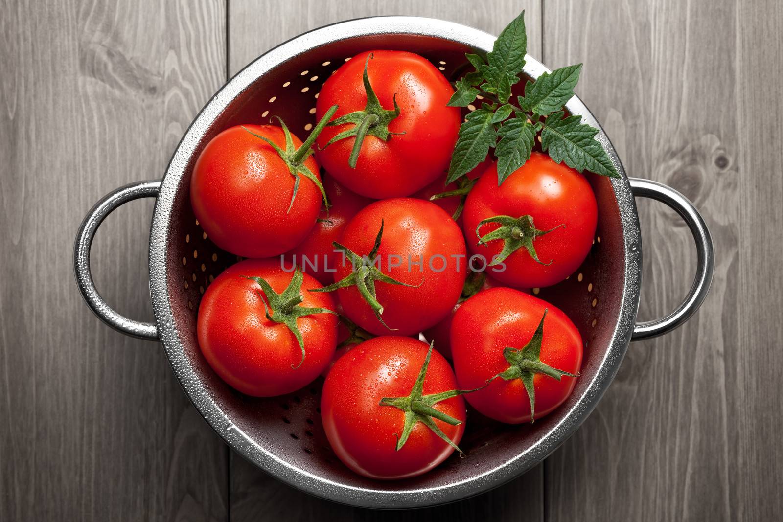 Fresh tomatoes with green leaf and waterdrops in colander on wooden background. Top view