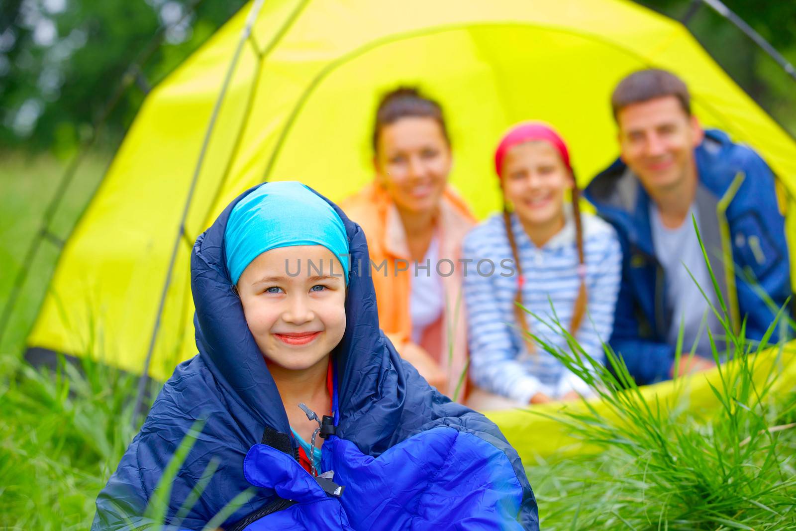 Young girl in a sleeping bag with her family near tent in camping on the nature