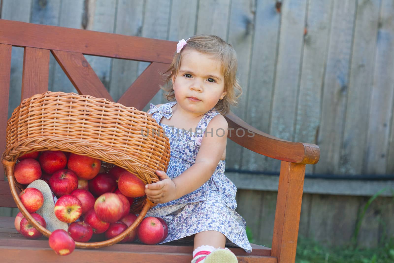 Little girl in a summer dress sits on a wooden shop with apples