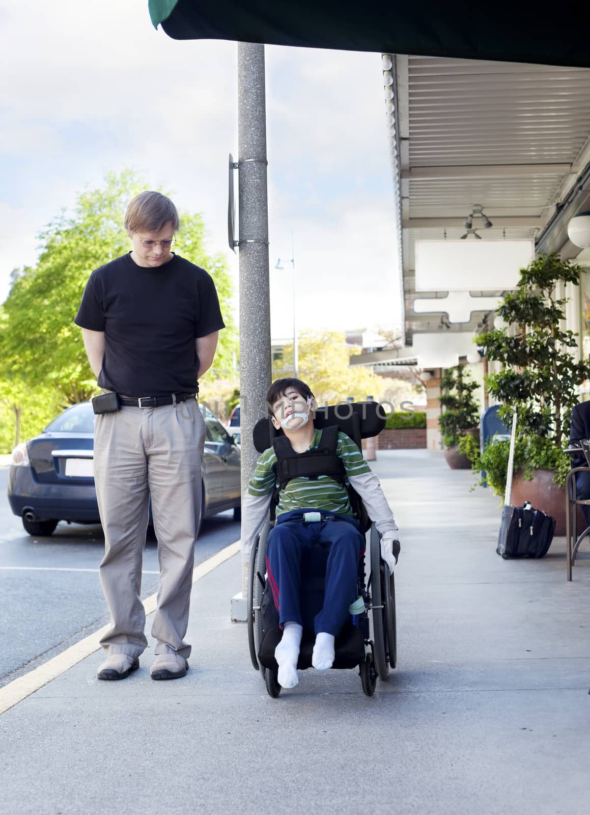 Father walking next to disabled son in wheelchair through town by jarenwicklund