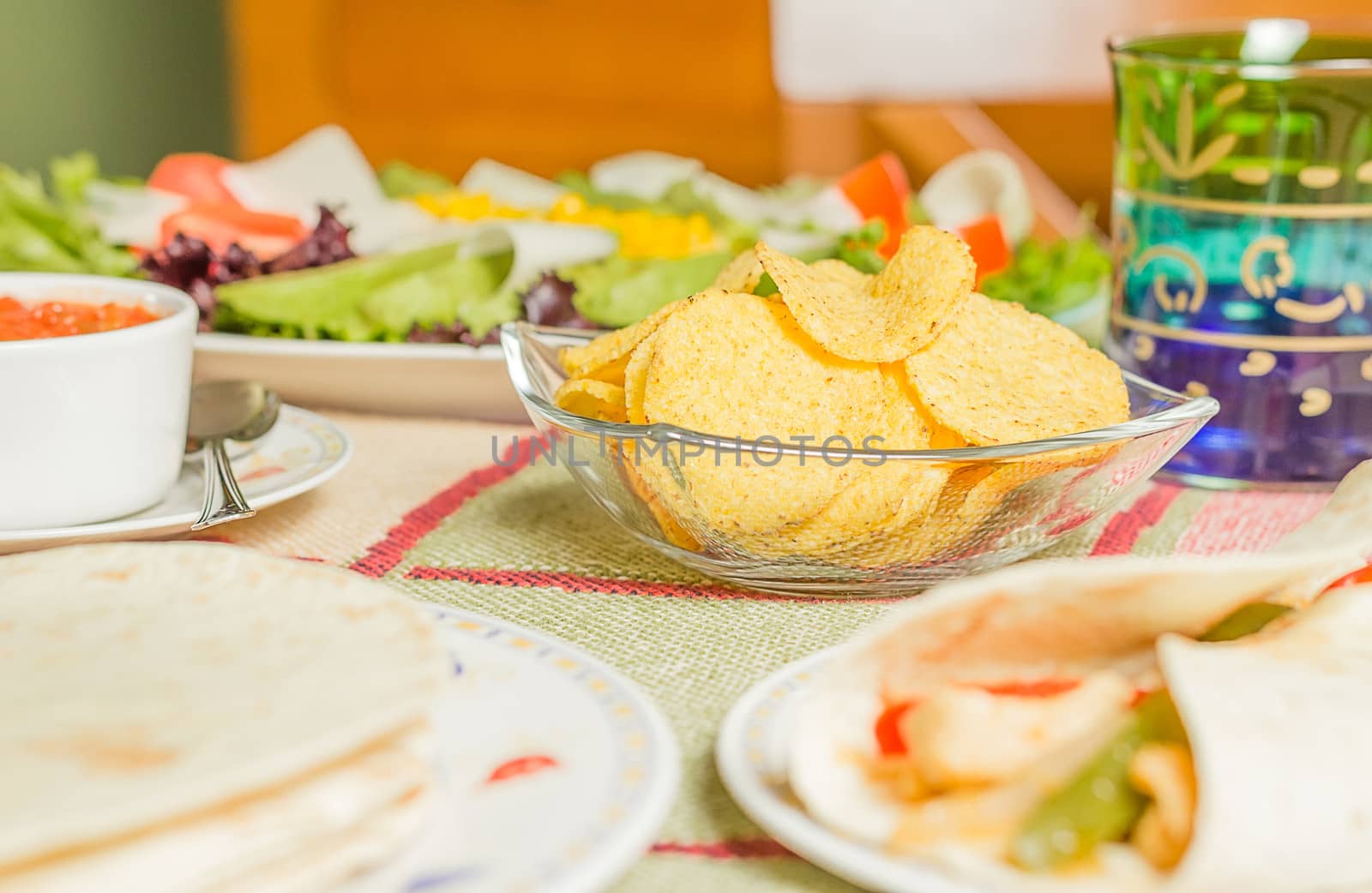 Closeup of traditional mexican food in a table, with  a bowl of nachos, spicy sauce, a plate of chicken fajita, tortillas and fresh salad
