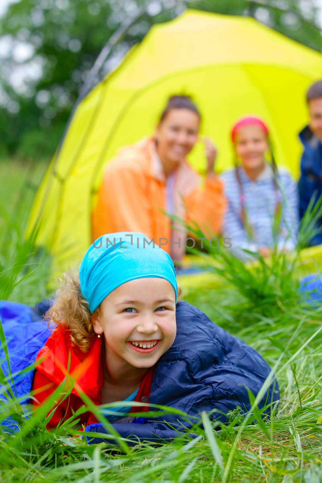 Young girl in a sleeping bag with her family near tent in camping on the nature. Vertical view