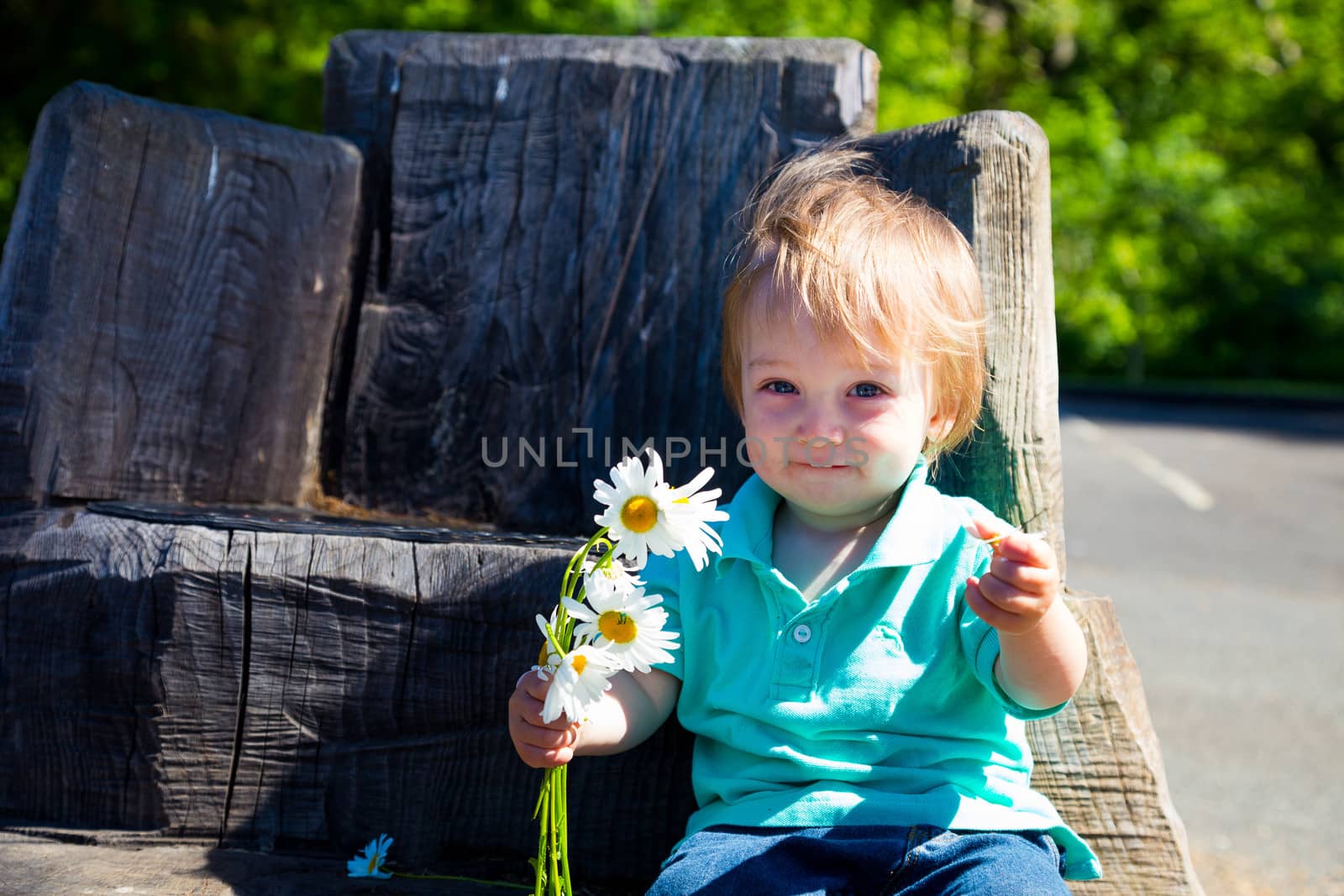 A young boy plays with some flowers while sitting on a hand carved seat made from an old stump.