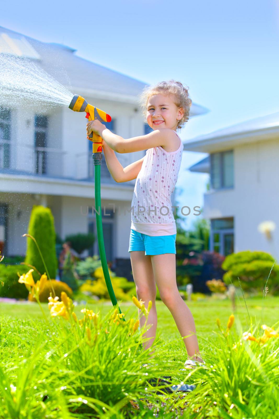 Little gardener girl watering flowers on the lawn near cottage. Vertical view