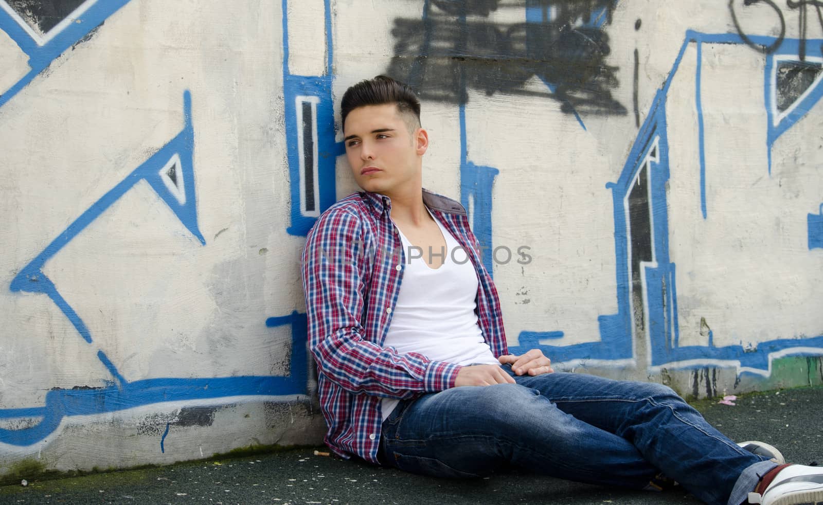 Handsome young man sitting on ground against colorful graffiti covered wall