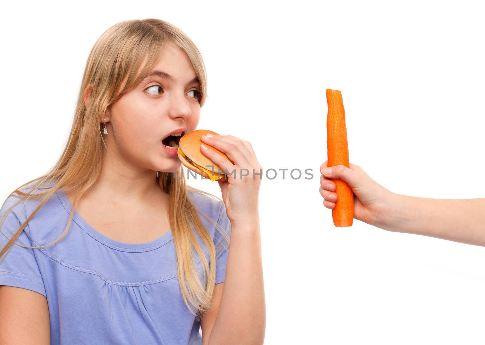 Young female eating a cheeseburger looking at a hand holding a carrot. Isolated on white.