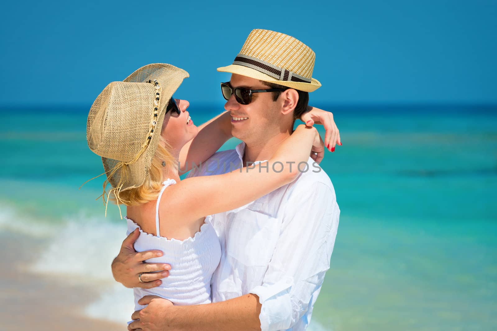 Happy embracing young couple in sunglasses, hats and white dress on a tropical beach with blue sea on background