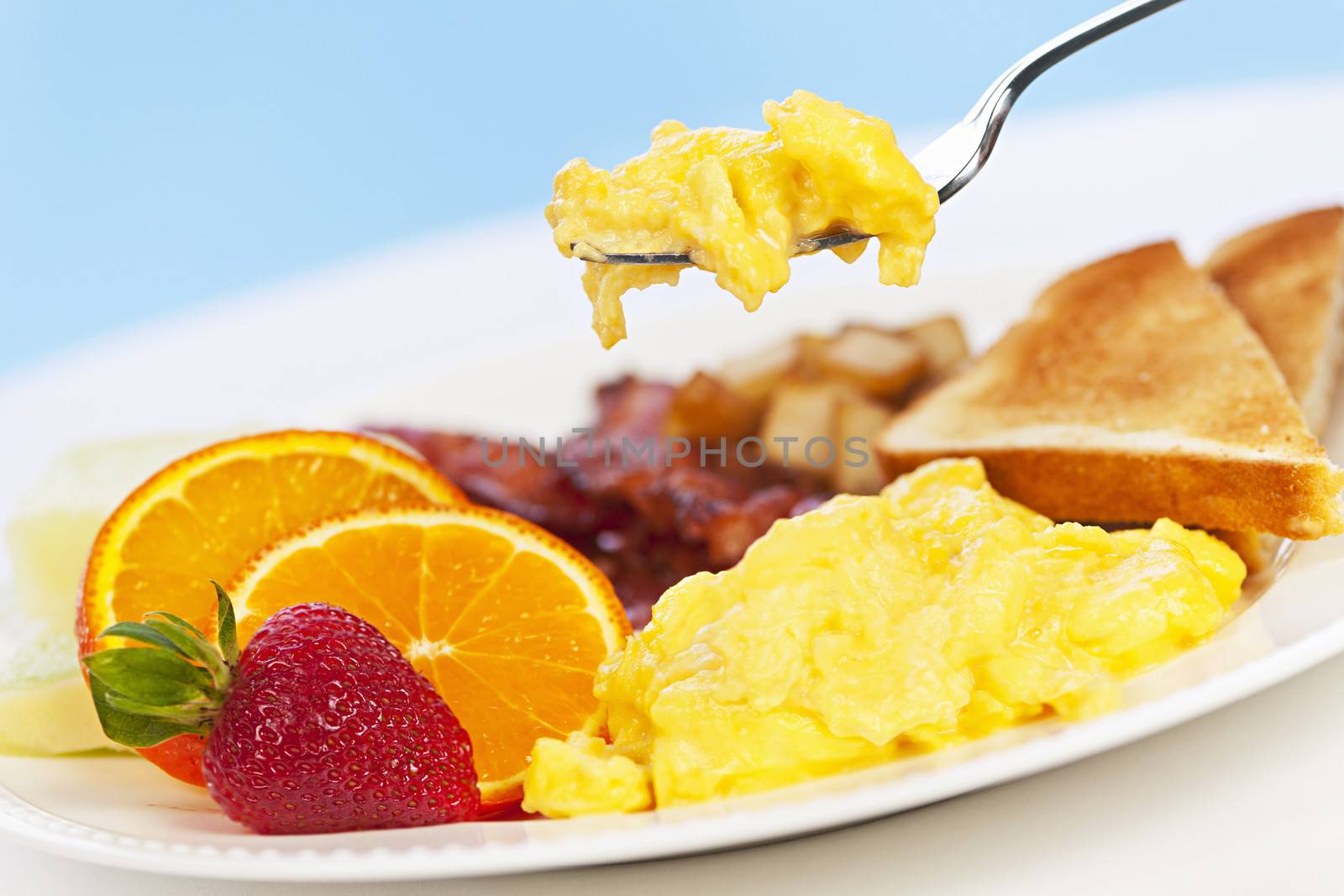 Scrambled eggs on a fork above breakfast plate with fruits toast and bacon
