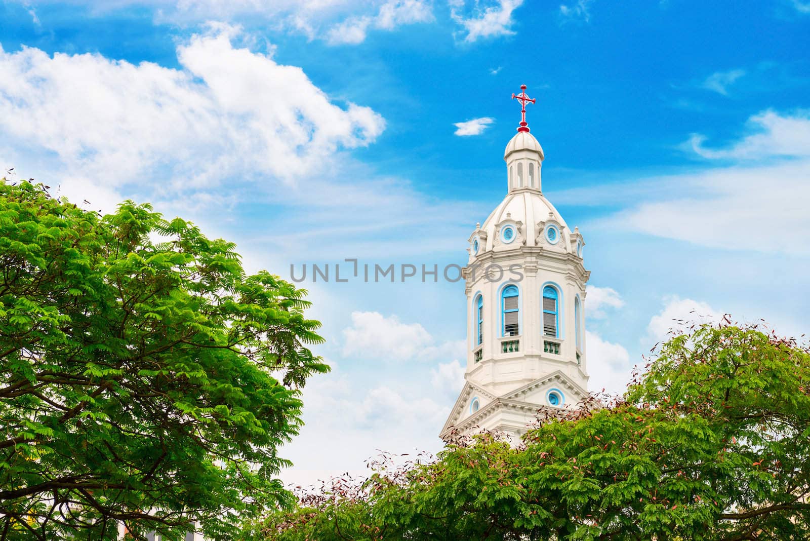 White church spire with trees and cloudy blue sky. Church of the Sacred Heart in Singapore.